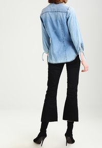 Camicia in denim azzurro chiaro con maniche lunghe e dettagli con fiocco, abbinata a pantaloni neri a campana e scarpe col tacco nere.