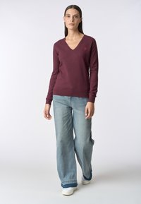 Maglione bordeaux a scollo a V in tessuto morbido, abbinato a jeans a gamba larga blu chiaro. Il maglione presenta un piccolo logo sul petto e maniche lunghe.