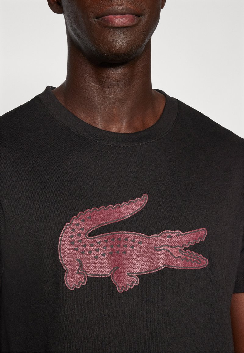 T-shirt noir à manches courtes, arborant un logo crocodile maroon texturé sur la poitrine. Tissu lisse avec un col ras du cou.