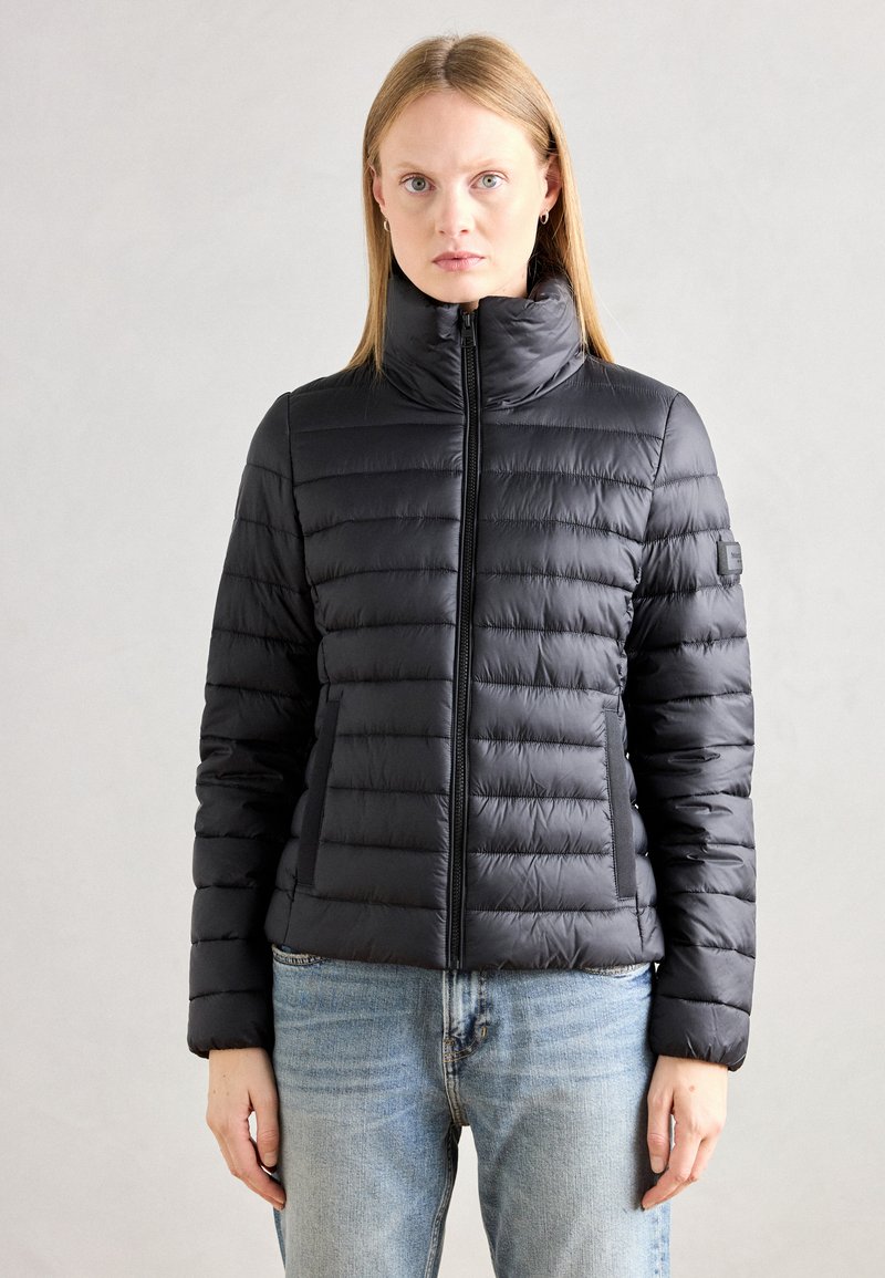 Doudoune noire avec un col haut, un motif matelassé horizontal et une fermeture éclair. Elle dispose de poches latérales et d'un patch logo sur la manche.