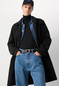Manteau long noir, veste en denim, col roulé noir, jeans bleu clair, ceinture noire et casquette. Style simple en couches avec des textures variées.