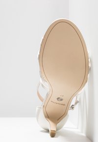 Semelle d'une chaussure de soirée blanche à talons hauts montrant le nom de la marque, la taille 37 et un petit élément décoratif en métal.