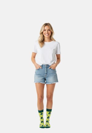 Femme souriante aux cheveux blonds, les mains dans les poches, portant un t-shirt blanc, un short en jean et des chaussettes jaunes avec un motif avocat.
