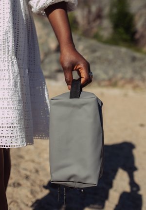 Mørkhudet hånd holder grå rektangulær veske ved svart stropp, iført hvit hullsømkjole, sandete utebakgrunn med steiner.