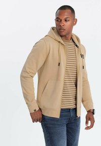 Sudadera beige con cremallera y cordón, bolsillos frontales y detalles en negro. Llevada sobre una camiseta de rayas, combinada con vaqueros azules. Textura suave.