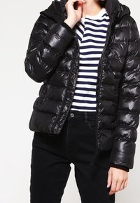 Osoba nosząca błyszczącą czarną kurtkę puchową na czarno-białą w paski koszulę i czarne spodnie, poprawiająca kołnierz kurtki.