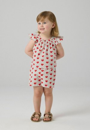 Toddler girl with light hair wearing cream outfit with red hearts and tan sandals, standing with hands behind back, smiling and looking right.