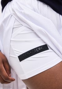 Κοντινό πλάνο ενός ατόμου που σηκώνει μια λευκή φούστα με τρύπες για να αποκαλύψει λευκά σορτς με μαύρο λογότυπο JLindeberg στο μηρό.