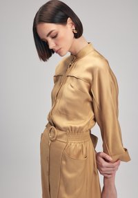 Vestido camisero de satén beige con cuello, cierre frontal de botones, dos bolsillos en el pecho y cintura elástica con cinturón y hebilla circular.