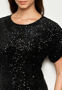 Černý top vyrobený z texturované látky s flitry, má kulatý výstřih a krátké rukávy. Design má těsný střih s viditelnými ozdobami.