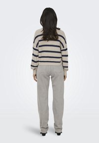 Femme aux longs cheveux foncés portant un pull beige à rayures bleu marine et un jean gris clair, debout de dos sur un fond blanc.