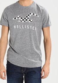 Šedé bavlněné tričko s černobílým kostkovaným potiskem racka, s nápisem "HOLLISTER" níže, v kombinaci s tmavými džíny.