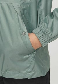 Mano nella tasca di una giacca verde chiaro con trama, polsino elastico e dettagli con bottoni automatici, indossata sopra pantaloni neri.