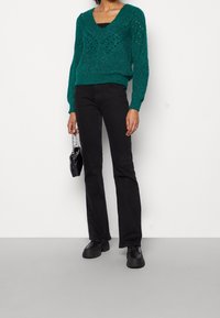 Pull tricoté couleur sarcelle avec un col en V et un motif en ajouré, associé à un jean noir évasé et des chaussures noires à lacets. Petit sac à main noir.