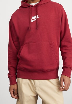 Mężczyzna w bordowej bluzie z kapturem Nike z przednią kieszenią i beżowych spodniach, stojący na jednolitym białym tle.