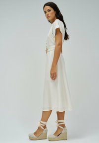 Vestido midi blanco con mangas cortas, cuerpo ajustado y detalles de botones dorados. Combinado con sandalias de cuña beige con ataduras en el tobillo.
