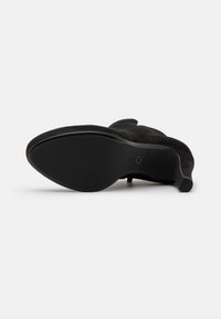 Botte à talon haut en suède noir avec une semelle lisse, un bout pointu et un talon effilé et courbé, mettant en valeur une finition mate et des détails de couture subtils.