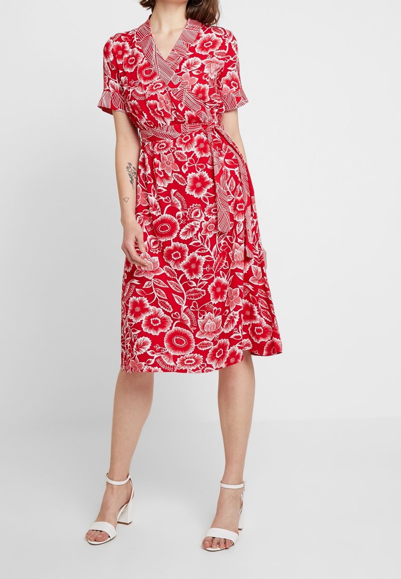 Femme portant une robe portefeuille rouge à motifs floraux blancs et des sandales à talons blocs à bride cheville blanches, debout devant un fond uni.