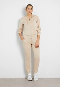 Bež pulover s kapuco z zadrgo in vrvico ter logotipom, v kombinaciji z ujemajočimi se zoženimi tekaškimi hlačami. Gladek material, rebrasti manšeti in sproščen kroj. Bele superge.