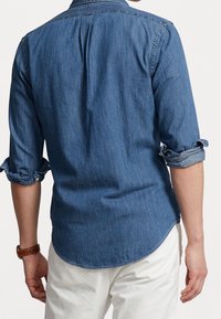Denimskjorta i mörkblått, med långärmar som är uppvikta. Har en klassisk krage och rundad nederkant. Bakom finns subtila veck för en skräddarsydd passform.