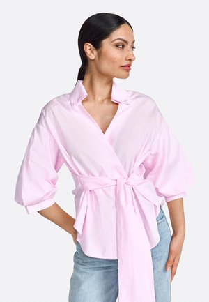 Rosa Wickelbluse mit tiefem V-Ausschnitt, übergroßen Ärmeln und gebundenem Taillenband. Hergestellt aus Baumwollstoff mit glatter Textur.