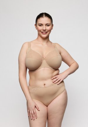 Lachende vrouw met donker haar in een nude kanten bh en bijpassende taillehoog slip staat tegen een effen witte achtergrond.