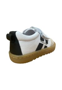 Zapatilla de cuero blanca y negra con acabado texturizado, suela de goma y correa de Velcro. Presenta un pequeño tirador en el talón para ponerla y quitarla con facilidad.