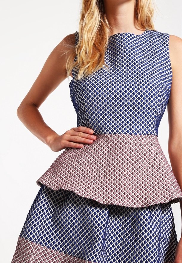 Femme portant une robe peplum sans manches bleue et rose texturée avec un motif géométrique, main posée sur sa taille.