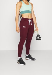 Sportovní outfit zahrnuje mintově zelenou a tyrkysovou sportovní podprsenku Nike, bordó tepláky s bílým šněrováním a černé tenisky s bílými detaily.