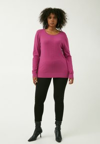 Maglione rosa a maniche lunghe con motivo a maglia lungo il centro, abbinato a pantaloni neri a vita alta e stivale con tacco neri, su uno sfondo neutro.