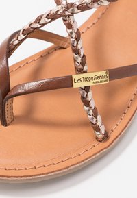Sandale en cuir marron avec des accents métalliques tressés, dotée d'une plaque logo en ton or et d'une semelle intérieure lisse en cuir beige.