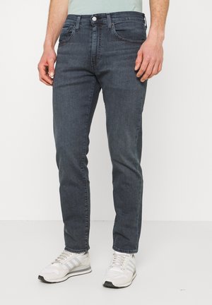 Homme portant un jean slim gris foncé et des baskets blanches, debout sur un fond blanc uni.