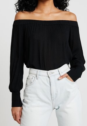 Femme portant un haut noir à manches longues et épaules dénudées, et un jean bleu clair taille haute, avec une main dans la poche.