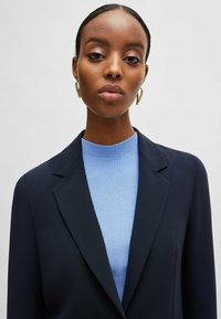 Marinblå kavaj med slag över en ljusblå ribbad tröja; guldörhängen som accentsmycken; slätt tyg, skräddarsydd passform.