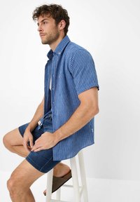Camicia a maniche corte a righe blu e bianche con colletto button-down. Abbinata a pantaloni corti di denim scuro e sandali marroni senza lacci.