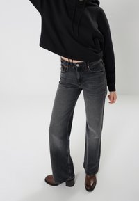 Vaqueros de denim negros de pierna ancha con un acabado desgastado, combinados con una sudadera negra corta. Botines de tacón marrones completan el look.