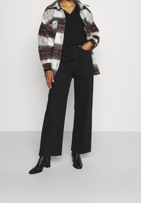 Schwarze weite Jeans kombiniert mit einem schwarzen Stricktop unter einer rot-weißen und grauen karierten oversized Hemdjacke. Schwarze Stiefeletten.