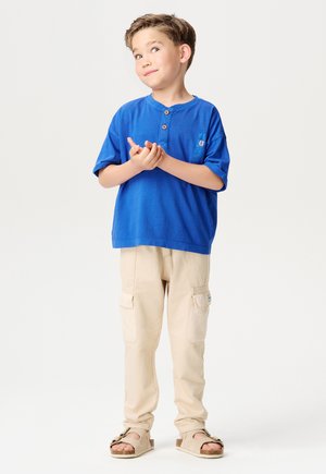Blaues T-Shirt mit kurzen Ärmeln und Knopfleiste, kombiniert mit beigen Cargo-Hosen mit Seitentaschen und braunen Sandalen mit Schnallen.
