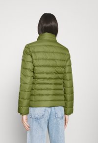 Oliven grøn pufferjakke med høj krave, vertikal quiltet tekstur og lange ærmer. Båret over lyseblå jeans.
