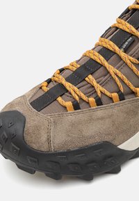 Chaussure en suede marron et textile avec semelle en caoutchouc noir ; présente des lacets orange et noirs, un embout renforcé et une texture de grip au fond.