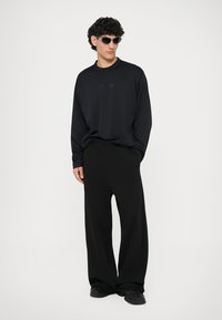 Long-sleeve black t-shirt with subtle logo, paired with wide-leg black trousers, and dark sunglasses, featuring a smooth fabric texture.