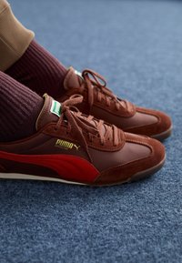 Bruna mocka- och lädersneakers med röda accenter, guldlogotyp och bruna snörning. Bärs tillsammans med ribbade bordeauxfärgade strumpor på en blå texturerad matta.