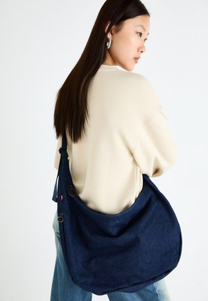 Dark denim shoulder bag with a soft, slouchy shape, adjustable strap, and zipper details featuring metallic accents. Logo visible on the front.
