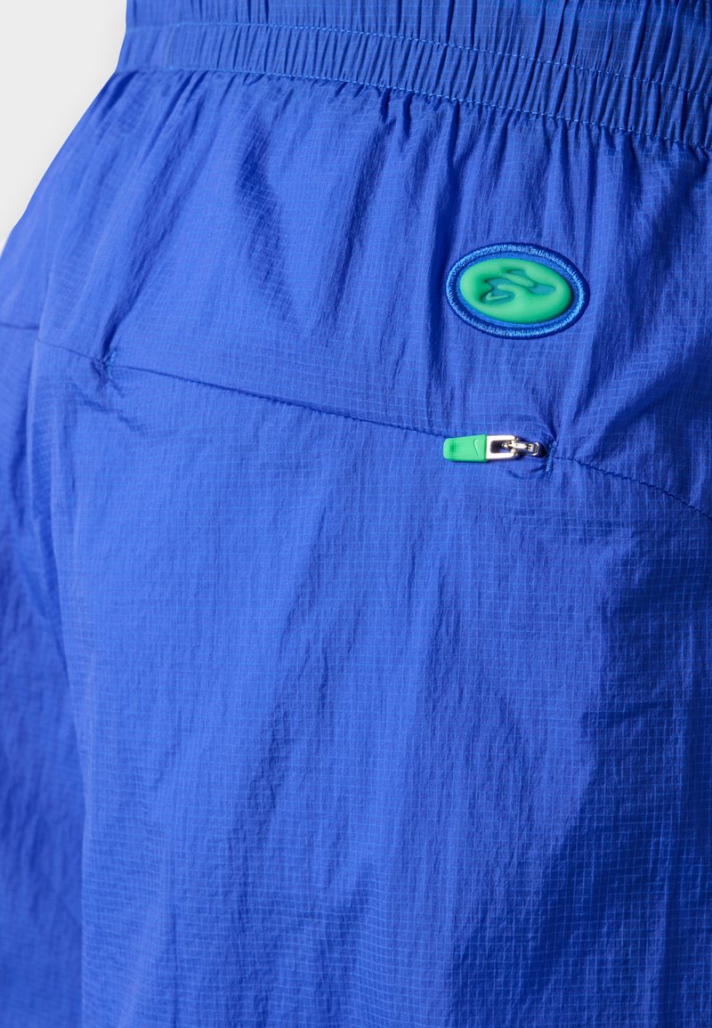 Blaue leichte Shorts aus strukturiertem Stoff mit einem elastischen Bund, einer mit Reißverschluss versehenen Tasche und einem grünen Logo-Patch.