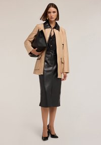 Beige vnější bunda s černým límcem, ve spojení s černou koženou tužkovou sukní, držící velkou černou kabelku, doplněné o špičaté podpatky.