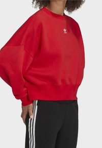 Rode sweatshirt gemaakt van zacht materiaal, met een ronde hals, verlaagde schouders en een klein wit logo op de borst.