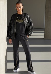 Čierna kožená bomber bunda cez čierne tričko so zlatým logom, spárovaná s čiernymi teplákovými nohavicami so zlatými zipsami a detailmi. Biele tenisky.