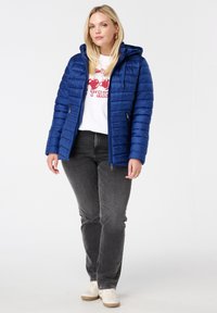 Blauwe pufferjack met een capuchon, horizontaal gestikt ontwerp, voorkruipzakken, gecombineerd met een wit T-shirt en grijze jeans. Gelaagd materiaal.