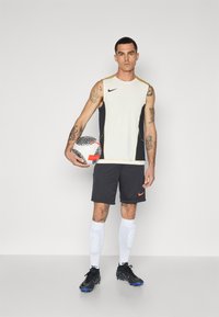 Nike športna majica brez rokavov z kremasto sprednjo stranjo in temnosivimi stranmi, v kombinaciji s temnimi kratkimi hlačami, belimi nogavicami in črnimi nogometnimi čevlji, drži nogometno žogo.