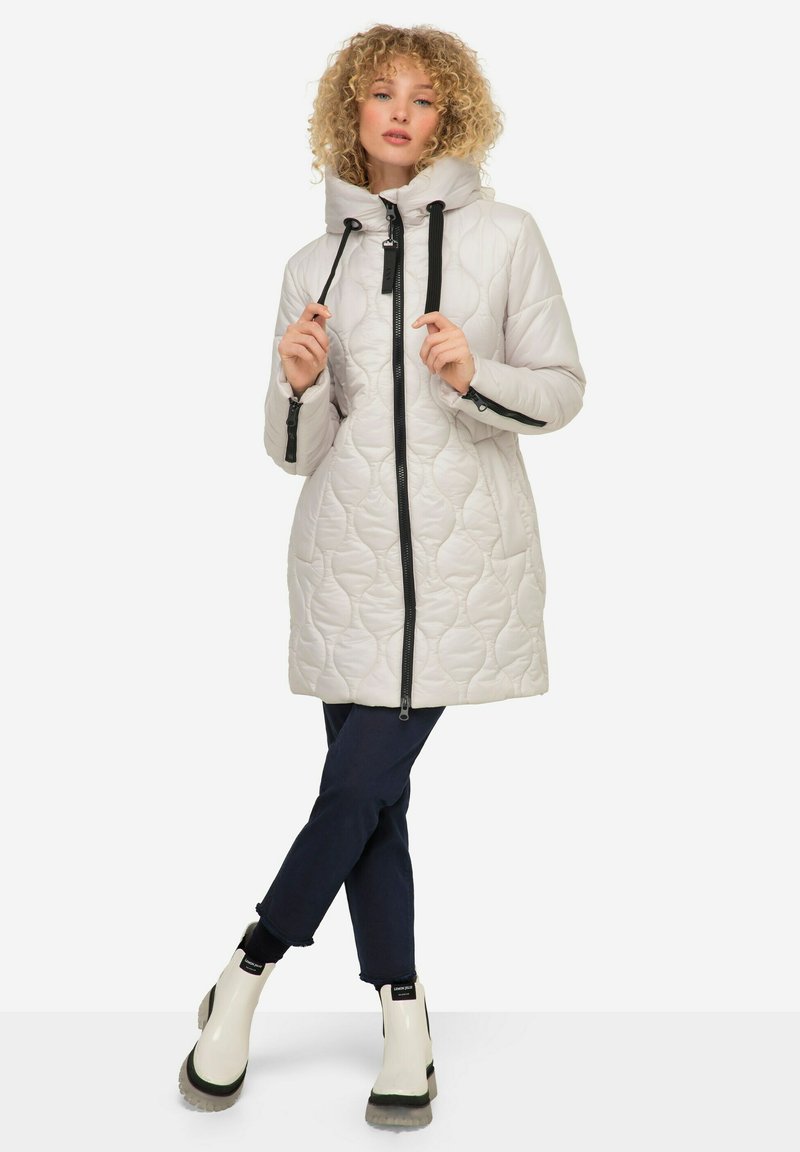 Manteau beige matelassé léger avec un col haut, une fermeture éclair noire et des cordons de serrage. Associé à un jean sombre et des bottines blanches avec des accents noirs.